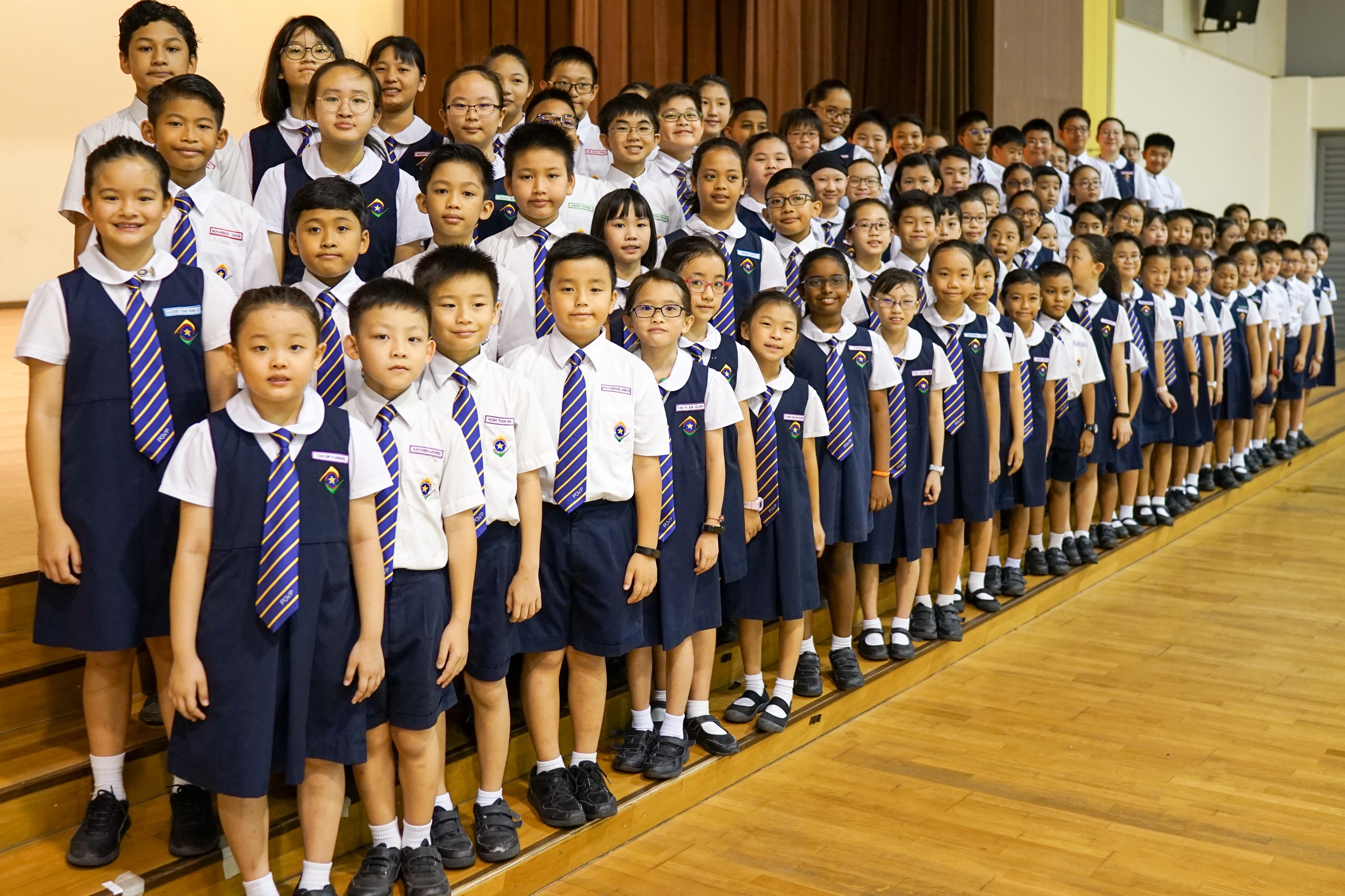 Large group of young students in navy and white uniforms with striped ties posing on tiered steps indoors.